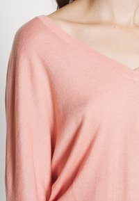 Jasnoróżowy sweter z dekoltem w kształcie litery V, wykonany z miękkiej dzianiny, charakteryzujący się luźnym krojem i ryżowym wykończeniem dekoltu, ukazujący delikatną fakturę.