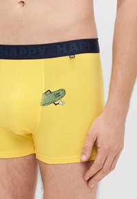 Sárga pamut boxernadrág tengerészkék elasztikus derékrésszel, amelyen a "HAPPY" felirat látható. Az elején egy zöld uborka figura grafikája található.
