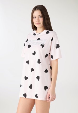 Giovane donna con capelli castani lunghi che indossa un vestito t-shirt oversize rosa chiaro con motivi a cuore neri su sfondo bianco.