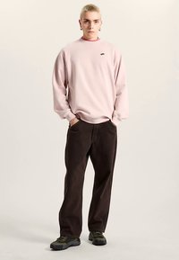 Sudadera de gran tamaño de color rosa claro con cuello redondo acanalado y un pequeño gráfico en el pecho, combinada con pantalones de corte holgado color marrón oscuro y zapatillas negras gruesas.