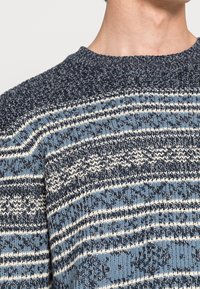 Pull en maille épaisse avec des rayures horizontales bleu marine, bleu clair et blanc sur le haut du torse et le cou d'une personne.