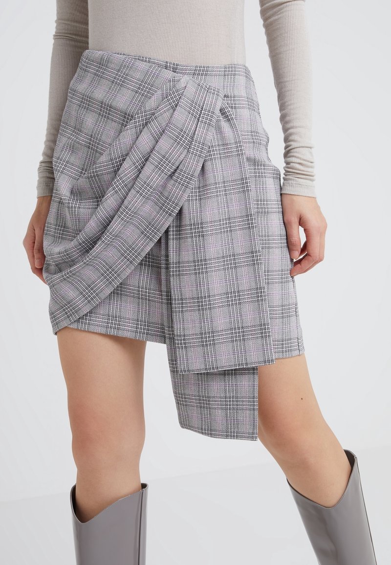 Femme portant une mini-jupe asymétrique à carreaux gris avec détail drapé, un top à manches longues beige et des bottes grises mi-mollet.