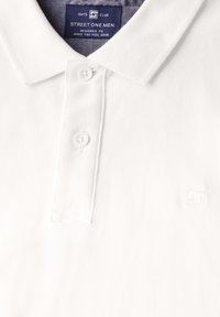 Bílá polo tričko vyrobené z texturované tkaniny, s tříknoflíkovou zápinkou a malým vyšitým logem na hrudi.