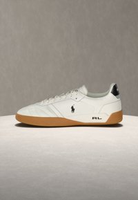 Λευκό sneaker χαμηλού ύψους με καφέ σόλα, μαύρο λογότυπο αλόγου και αναβάτη στο πλάι, μαύρη λωρίδα τακουνιού και γράμματα "RL" κοντά στη σόλα.