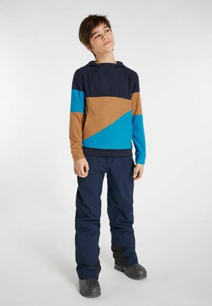Felpa con cappuccio a blocchi di colore blu navy, marrone e turchese; texture morbida; design casual; indossata con pantaloni scuri e stivali.