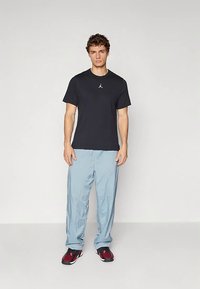 Melna īsroku T-krekls ar mazu sudrabotu logotipu, kombinēts ar gaiši zilām vaļīgām biksēm un melni sarkaniem sporta apaviem. Vienkāršs fons.