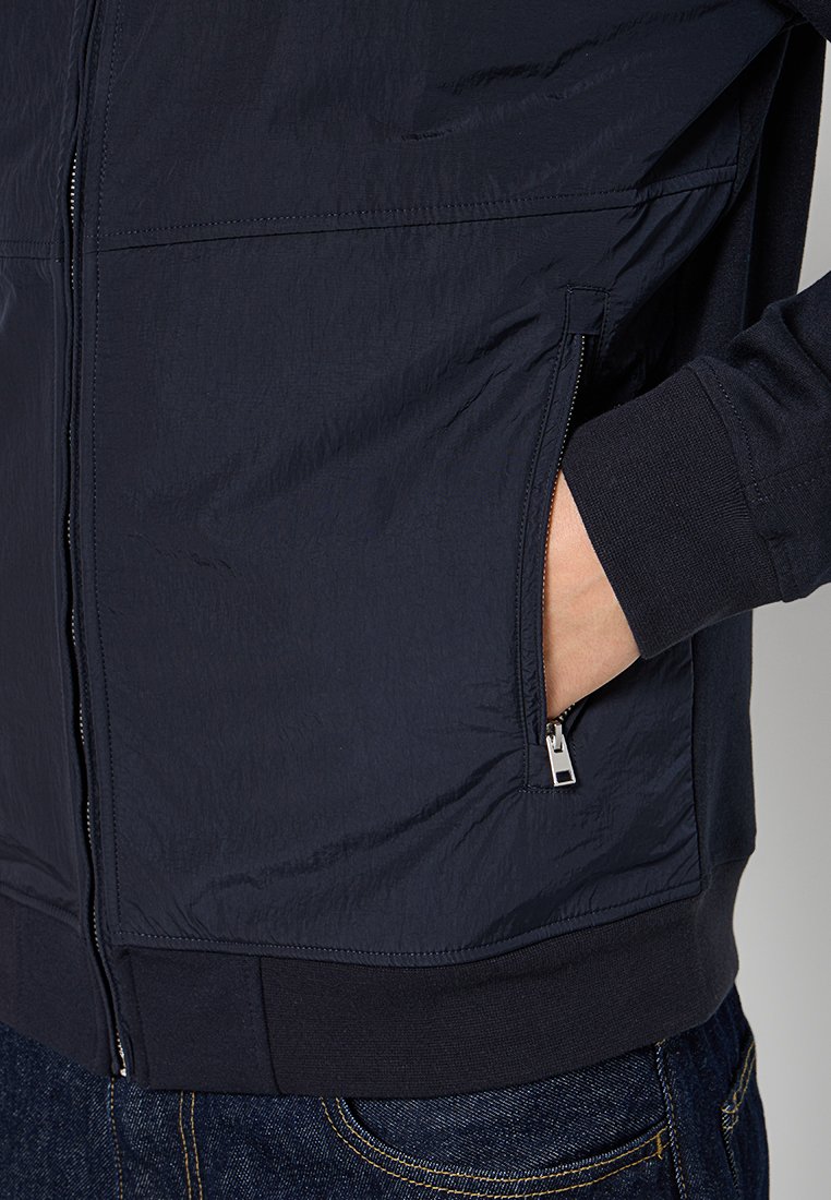 Veste légère navy avec poches zippées, tissu texturé et poignets côtelés. Conçue pour le confort et la fonctionnalité.