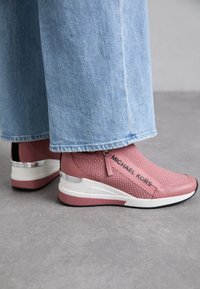Adidași textile roz cu suprafață din plasă, având un logo negru, talpă albă și accente metalice la călcâi, asortați cu blugi de denim de culoare albastru deschis.
