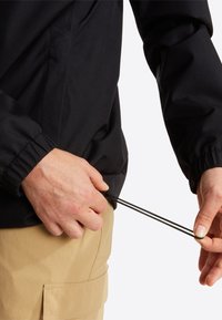 Χέρια που προσαρμόζουν το κορδόνι στο κάτω μέρος του μαύρου μπουφάν πάνω από μπεζ cargo παντελόνια σε λευκό φόντο.