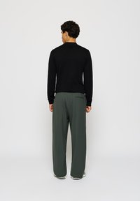 Zwart gebreid lange mouwen top met een kraag, gecombineerd met losse olijfgroene broek met een achterzak. Neutrale achtergrond.