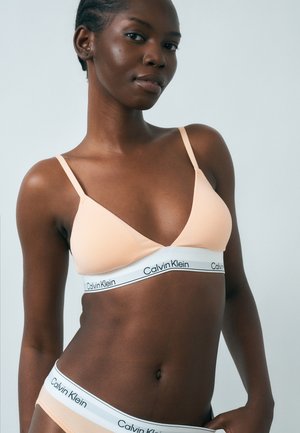 Femme portant un ensemble de sous-vêtements Calvin Klein couleur pêche clair avec des bandes élastiques blanches à logo, posant devant un fond gris clair uni.