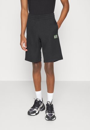 Personne portant un short noir avec le logo EA7, des baskets noires et blanches, des chaussettes blanches, debout avec une main dans la poche devant un fond uni.