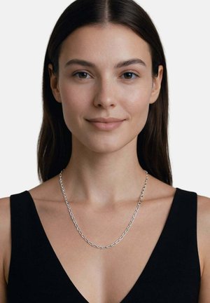 Giovane donna con capelli lisci e scuri che indossa una collana a catena argentata e una canotta nera senza maniche, che sorride dolcemente su uno sfondo semplice.