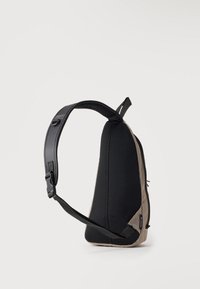 Kleiner beiger und schwarzer Sling-Rucksack mit gepolstertem Netzrücken und verstellbarem Einzelschulterriemen vor einfarbigem Hintergrund.