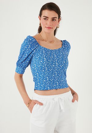 Mėlynas gėlių rašto crop top'as su pūstomis rankovėmis ir kvadratine iškirpte, turi mažą baltą gėlių raštą ir tekstūrinę medžiagą. Sukombinuotas su baltais keliais.
