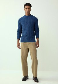 Granatowy dzianinowy sweter z teksturowanym wzorem diamentowym, z prążkowanym dekoltem i mankietami, zestawiony z jasnobeżowymi spodniami i czarnymi eleganckimi butami.