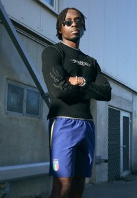 Camisola preta de mangas longas com detalhe de logotipo e mangas texturizadas, combinada com calções azuis com detalhes em amarelo e emblema da Itália.