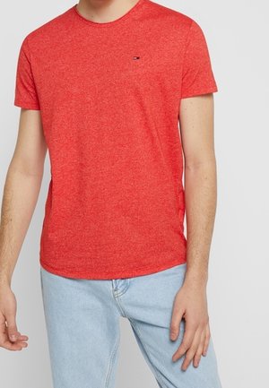Man draagt een effen rood T-shirt met korte mouwen en een klein logo op de borst, gecombineerd met lichtblauwe jeans, staand tegen een effen achtergrond.