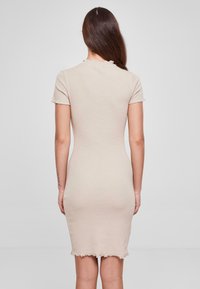 Robe bodycon beige à côtes avec des manches courtes, silhouette ajustée et ourlet brut. Présente un col rond subtil et une texture douce.