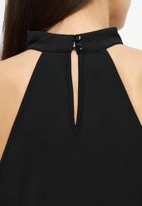 Πλάγια όψη μιας γυναίκας που φοράει μαύρο φόρεμα με λαιμόκοψη halter, στερεωμένο με δύο μαύρα κουμπιά και άνοιγμα σε σχήμα κλειδαρότρυπας στο άνω μέρος της πλάτης.