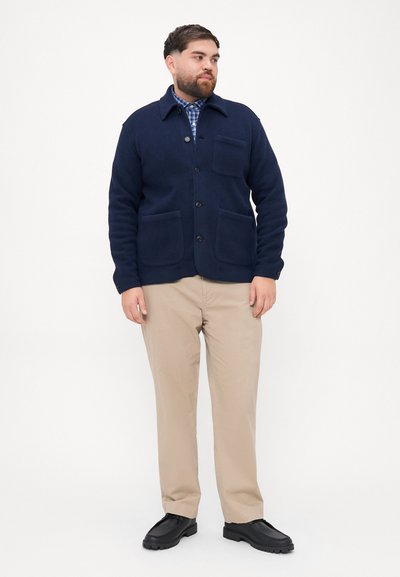 Μπλέ μάλλινη ζακέτα με γιακά, κουμπί και δύο μπροστινές τσέπες. Συνδυάζεται με μπεζ παντελόνια και μαύρα παπούτσια, φέρει καρό πουκάμισο.