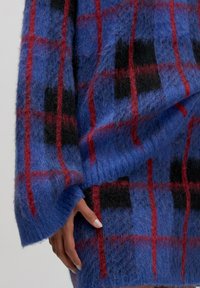 Hand in einem blauen, flauschigen Pullover mit rot-schwarzem Karomuster, die entspannt zur Seite liegt.
