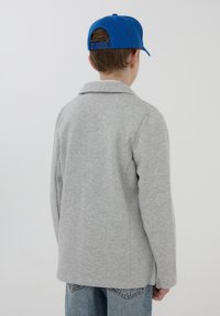 Felpa grigia con colletto, tessuto testurizzato e polsini con bottoni. Abbinata a un cappellino blu, vista da dietro, che mostra il design e la vestibilità casual.