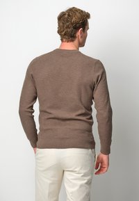 Maglione marrone strutturato con maniche lunghe e polsini a coste, abbinato a pantaloni di colore chiaro, visto da dietro.