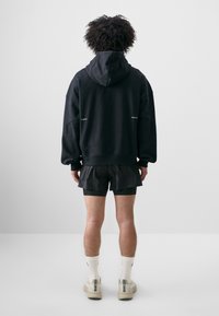Sort hættetrøje med oversized pasform, der har synlige syninger. Kombineret med sorte shorts og hvide sokker. Neutral baggrund.