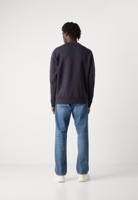 Βαμβακερή μπλούζα σε σκούρο ναυτικό χρώμα με στρογγυλή λαιμόκοψη, ραμμένες μανσέτες και στρίφωμα, συνδυασμένη με ανοιχτό γαλάζιο παντελόνι καμπάνα και μπεζ αθλητικά παπούτσια.