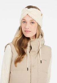 Bandeau en polaire beige avec un design torsadé, présentant une surface texturée. Le modèle porte un gilet matelassé beige, mettant en valeur le look superposé.