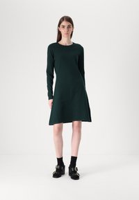 Σκούρο πράσινο φόρεμα μακρύ μανίκι με φουσκωτό φουστάνι, φτιαγμένο από μαλακό ύφασμα. Συνδυάζεται με μαύρα χοντρά τακούνια και κάλτσες μέχρι τον αστράγαλο.