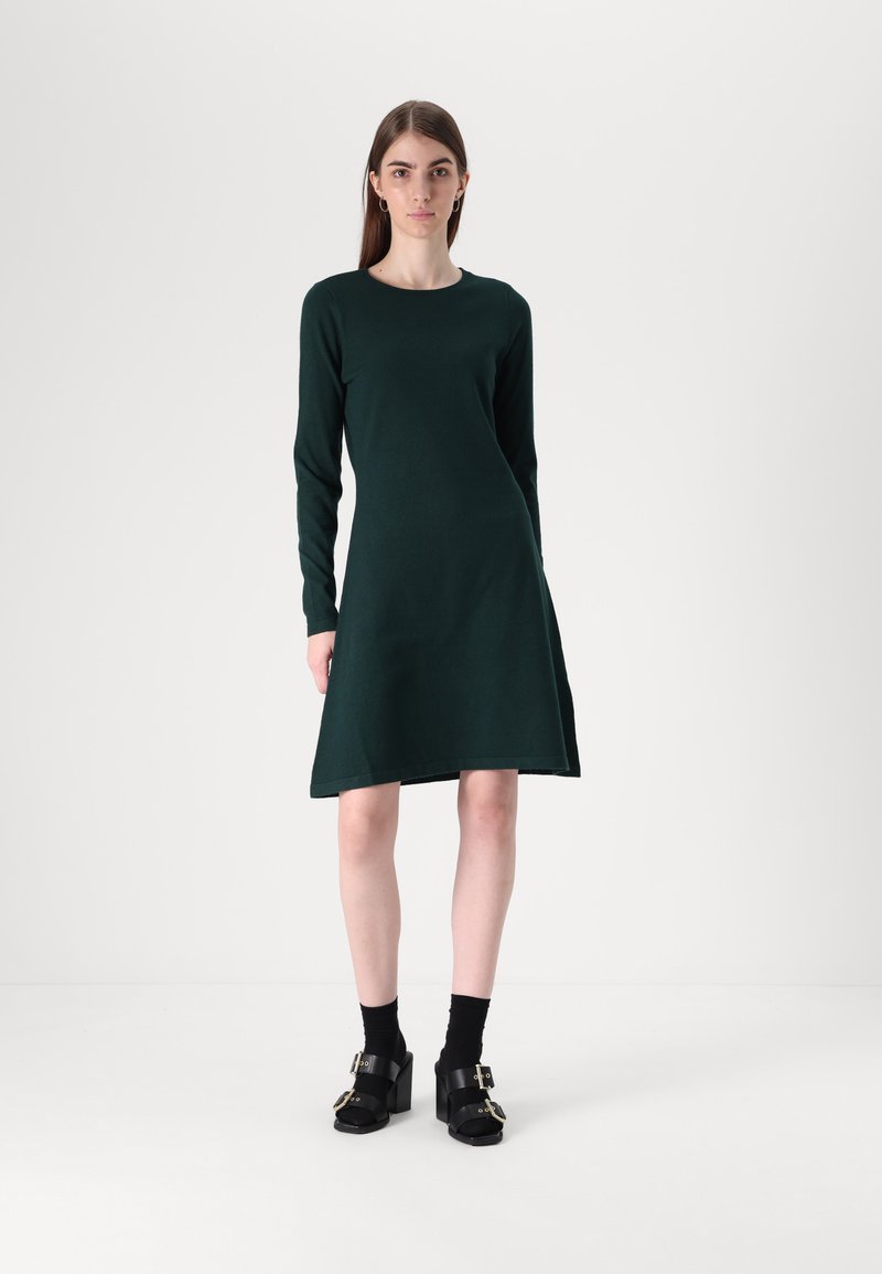 Σκούρο πράσινο φόρεμα μακρύ μανίκι με φουσκωτό φουστάνι, φτιαγμένο από μαλακό ύφασμα. Συνδυάζεται με μαύρα χοντρά τακούνια και κάλτσες μέχρι τον αστράγαλο.