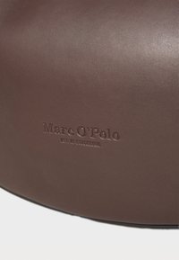 Borsa in pelle marrone con testo in rilievo "Marc O'Polo" e dettaglio "EST. IN STOCKHOLM". Texture liscia con un design curvo e minimalista.