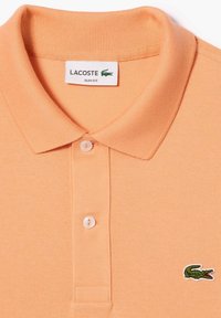 Polo shirt de corte slim en color durazno de Lacoste, con un tejido texturizado, cuello de canalé y tres botones blancos; cuenta con un logo de cocodrilo verde.