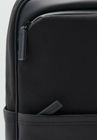 Mochila de tela negra con un acabado texturizado, que cuenta con un cierre de cremallera y un bolsillo exterior con una pequeña etiqueta. Acentos suaves en toda la mochila.