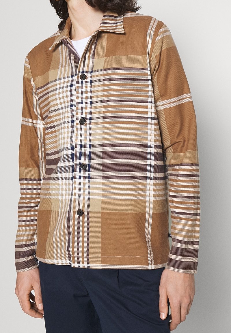 Hombre vestido con una camisa de cuadros marrón, beige y blanca, con cuello, combinada con pantalones oscuros, frente a un fondo claro y sencillo.