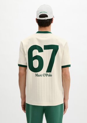 Homme portant une chemise crème à manches courtes avec bordure vert foncé et grand numéro 67, logo Marc O’Polo dans le dos, pantalon vert assorti, casquette blanche.