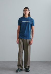 Tricou din bumbac de culoare bleu marin, cu "EMPORIO ARMANI" imprimat în alb, asortat cu pantaloni largi de culoare verde măsliniu și încălțăminte neagră.