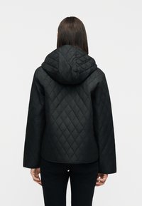 Γυναίκα που φαίνεται από πίσω να φοράει μαύρο καπιτονέ μπουφάν με κουκούλα και μακριά μανίκια, στέκεται μπροστά σε ένα απλό λευκό φόντο.