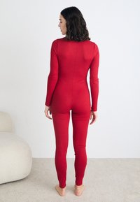 Piros bordás bodysuit hosszú ujjal, testhezálló fazonnal, sima textúrával és hátsó varrással. Meztelen lábakkal álló figura egy világos szőnyegen.