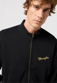 Chaqueta negra con cremallera, hecha de un tejido texturizado, destacando un logo dorado de "Wrangler". El alto cuello y la cremallera dorada añaden un contraste al diseño oscuro.