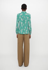 Green blouse with white floral and striped pattern, long sleeves, button-up back. Worn with high-waisted beige wide-leg trousers, featuring pockets.
