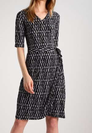 Woman wearing knee-length black wrap dress with white geometric triangle pattern and half sleeves, standing against plain background.