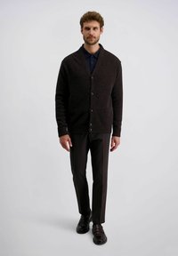 Man in een donkerbruine knopen cardigan, marineblauw shirt, donkere broek en zwarte leren schoenen die tegen een effen lichte achtergrond staat.