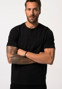 Černé polokošile s krátkým rukávem a tříknoflíkovou légou, s jemným logem. Model má viditelné tetování na rukou a stojí se založenýma rukama.