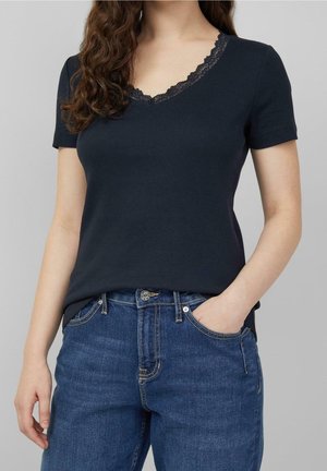 Vrouw draagt een zwart topje met korte mouwen en een kanten V-hals, gecombineerd met blauwe spijkerbroek, één hand in de zak, voor een grijze achtergrond.