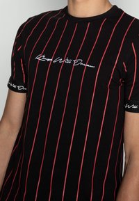 Czarna koszulka z pionowymi czerwonymi paskami, okrągłym dekoltem i krótkimi rękawami z kontrastowym logo na mankietach. Napis brzmi "Kings Will Dream."