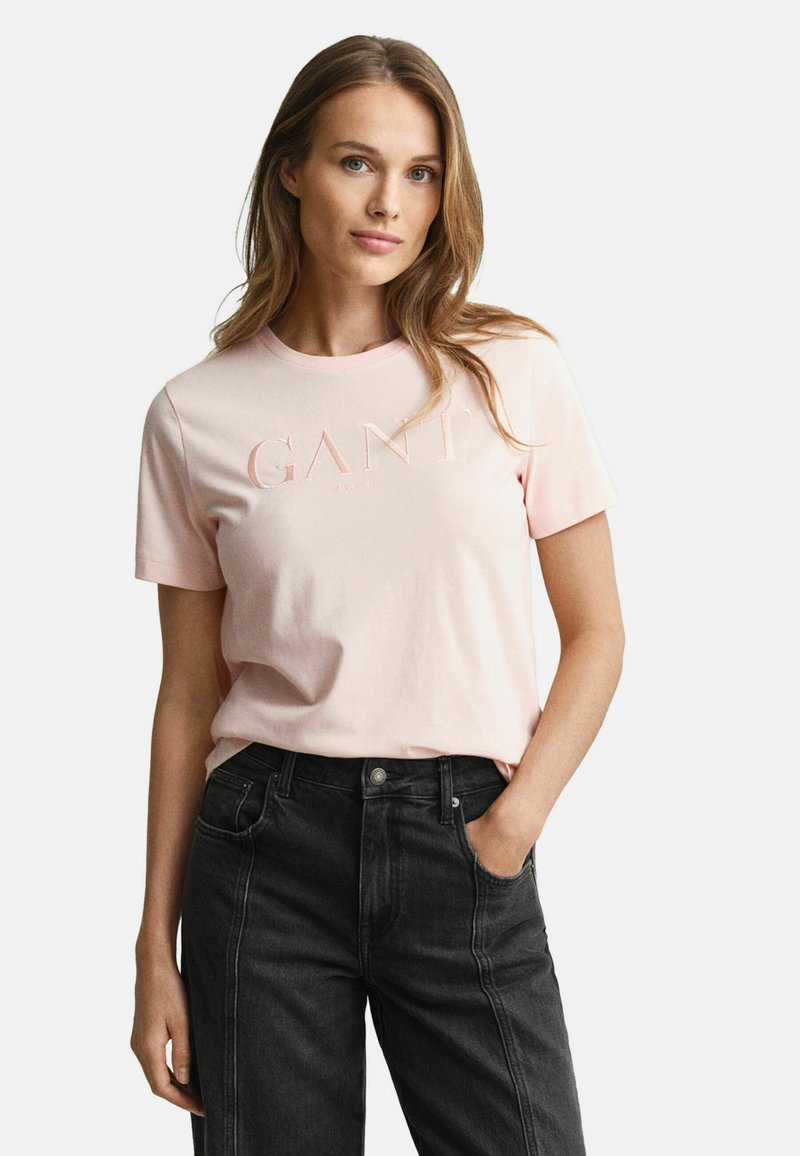 Helles rosa Baumwoll-T-Shirt mit Rundhalsausschnitt und kurzen Ärmeln, das ein geprägtes "GANT"-Logo auf der Vorderseite aufweist. Kombiniert mit schwarzen Jeans.