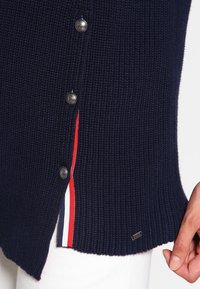 Námořnický pletený svetr s postranními knoflíky, s červenobílým pruhovaným detailem podél rozparku a malým kovovým logem.